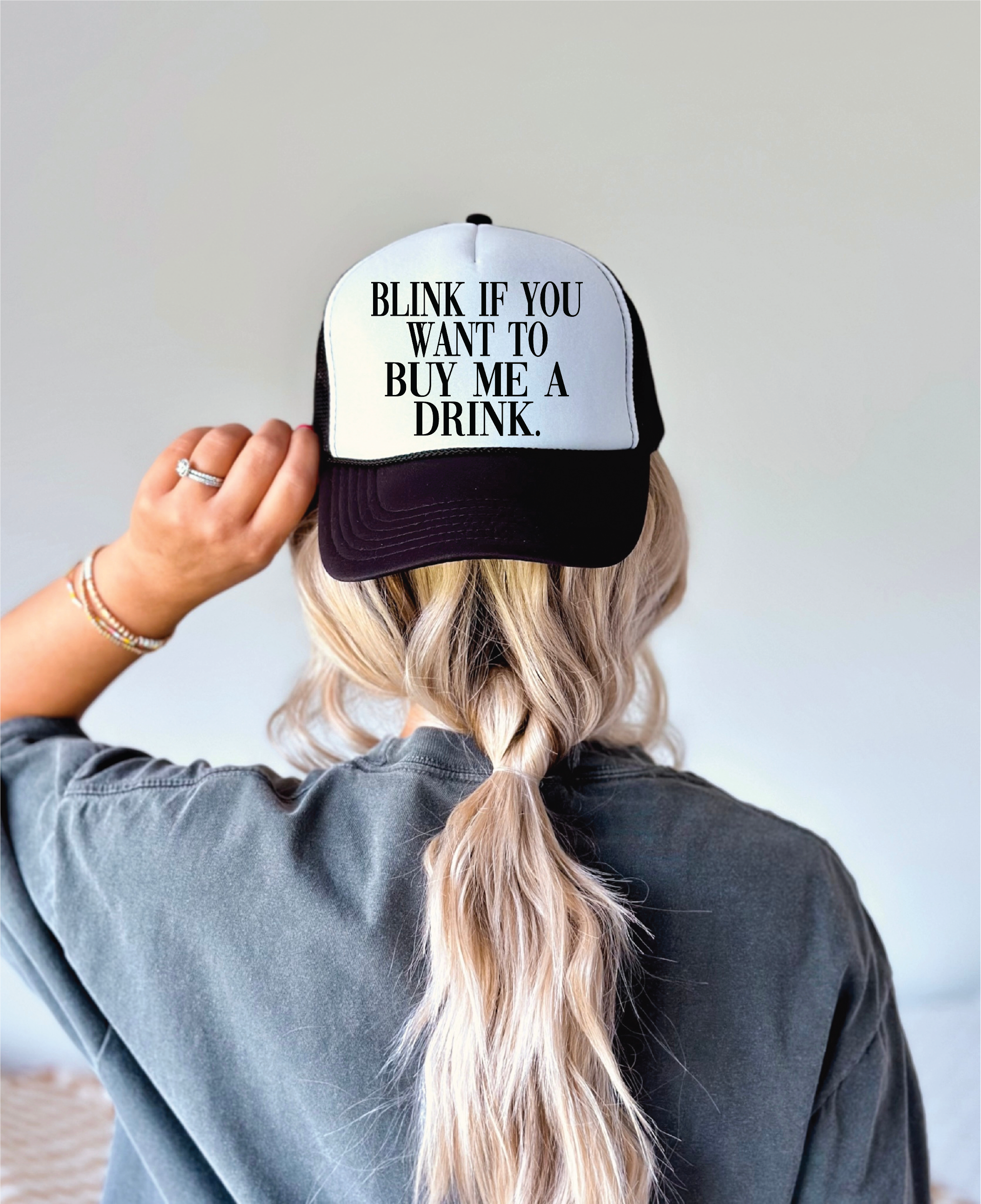 Person wearing a cap with text 'Blink if you want to buy me a drink' on a plain background