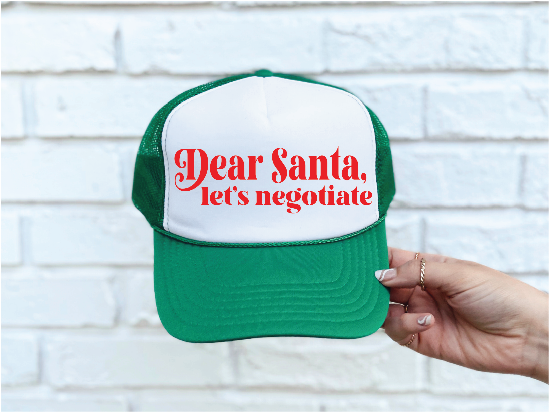 Green and white cap with 'Dear Santa, let's negotiate' text held against a white brick wall.