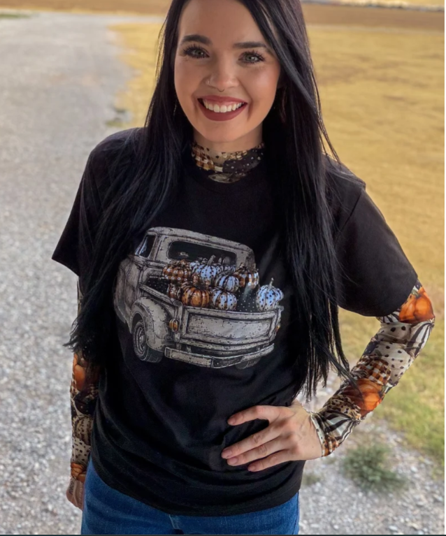 A woman smiling outdoors wearing a black graphic t-shirt with a vintage pickup truck carrying pumpkins, layered over a long-sleeve shirt with a pumpkin and plaid pattern on the sleeves.