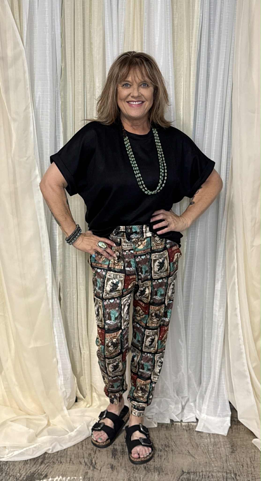 Woman wearing a black top and patterned pants standing in front of white curtains.