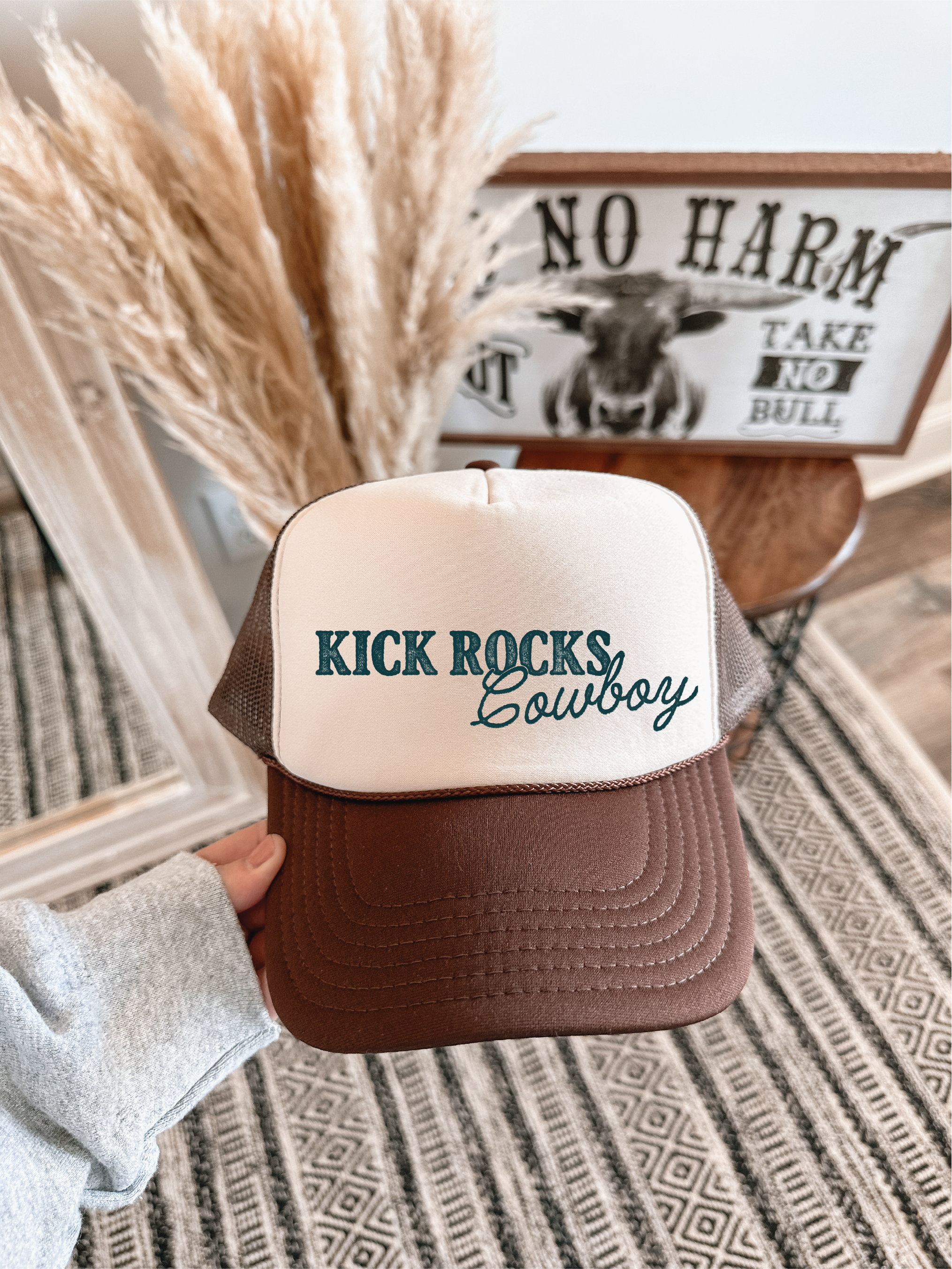 Brown and beige cap with 'Kick Rocks Cowboy' text held in front of a decorative wall with pampas grass and a framed picture.