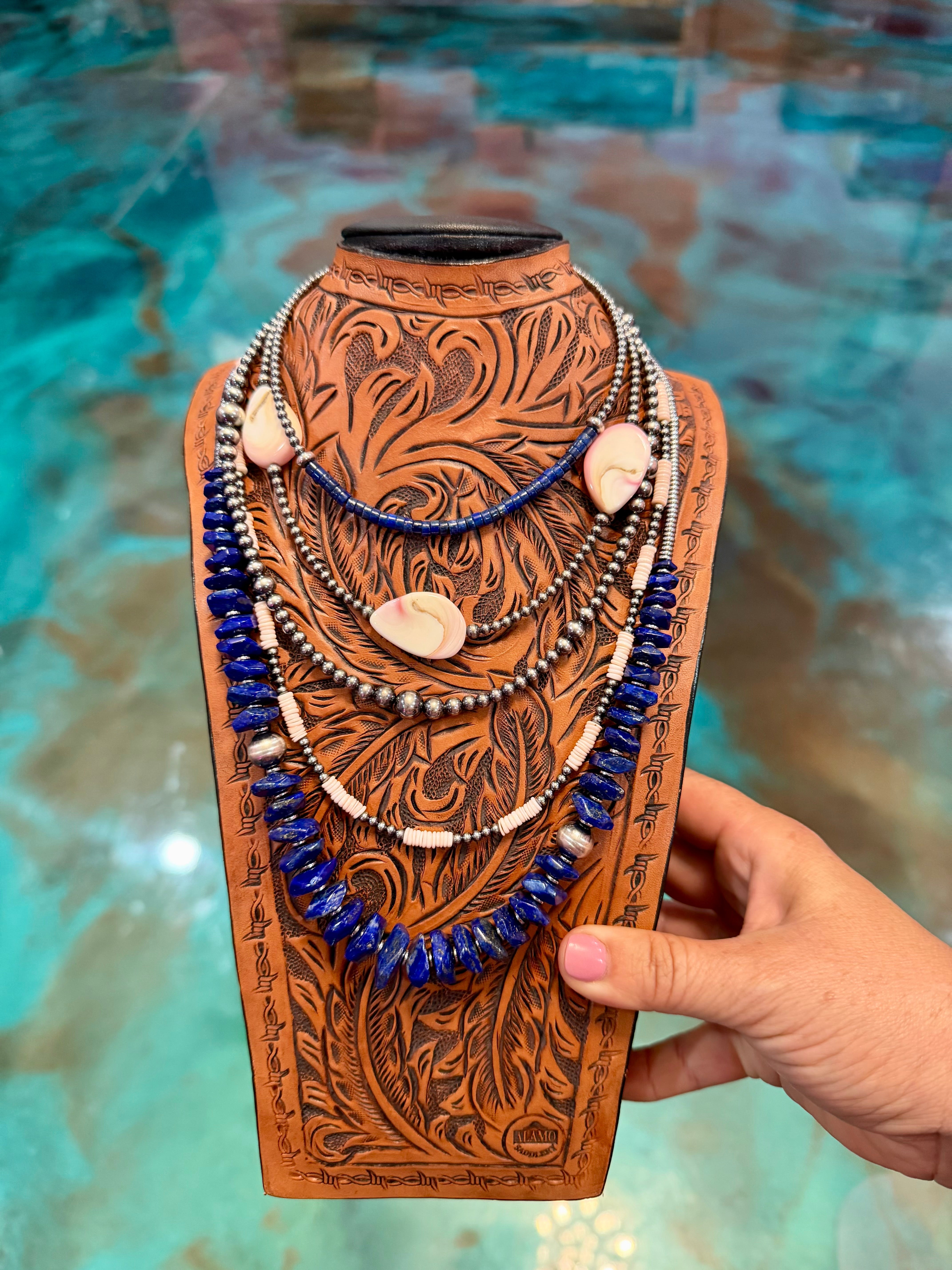Handmade leather necklace display with blue beads and shell elements against a textured turquoise background.