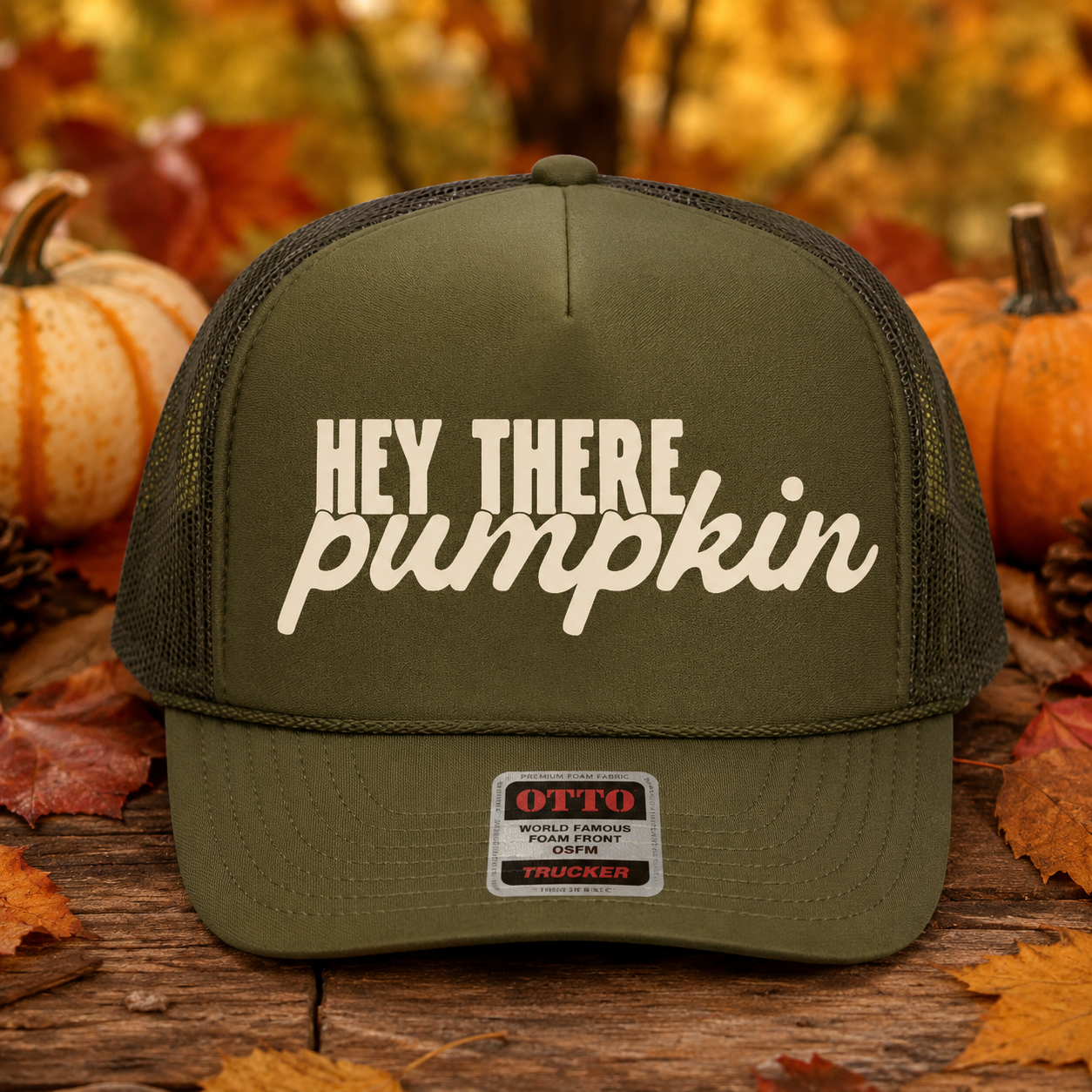 An olive green trucker hat with 'HEY THERE pumpkin' printed in white on the front, displayed on a rustic wooden surface with autumn leaves and pumpkins in the background.