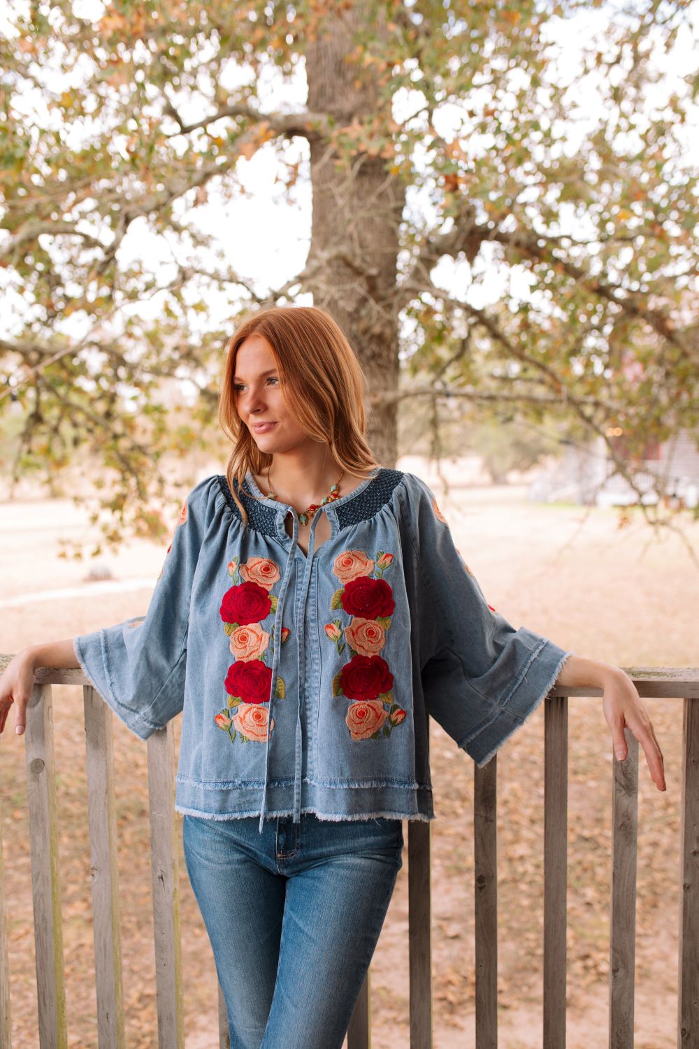 Woman wearing a denim jacket with floral embroidery, standing outdoors near trees.