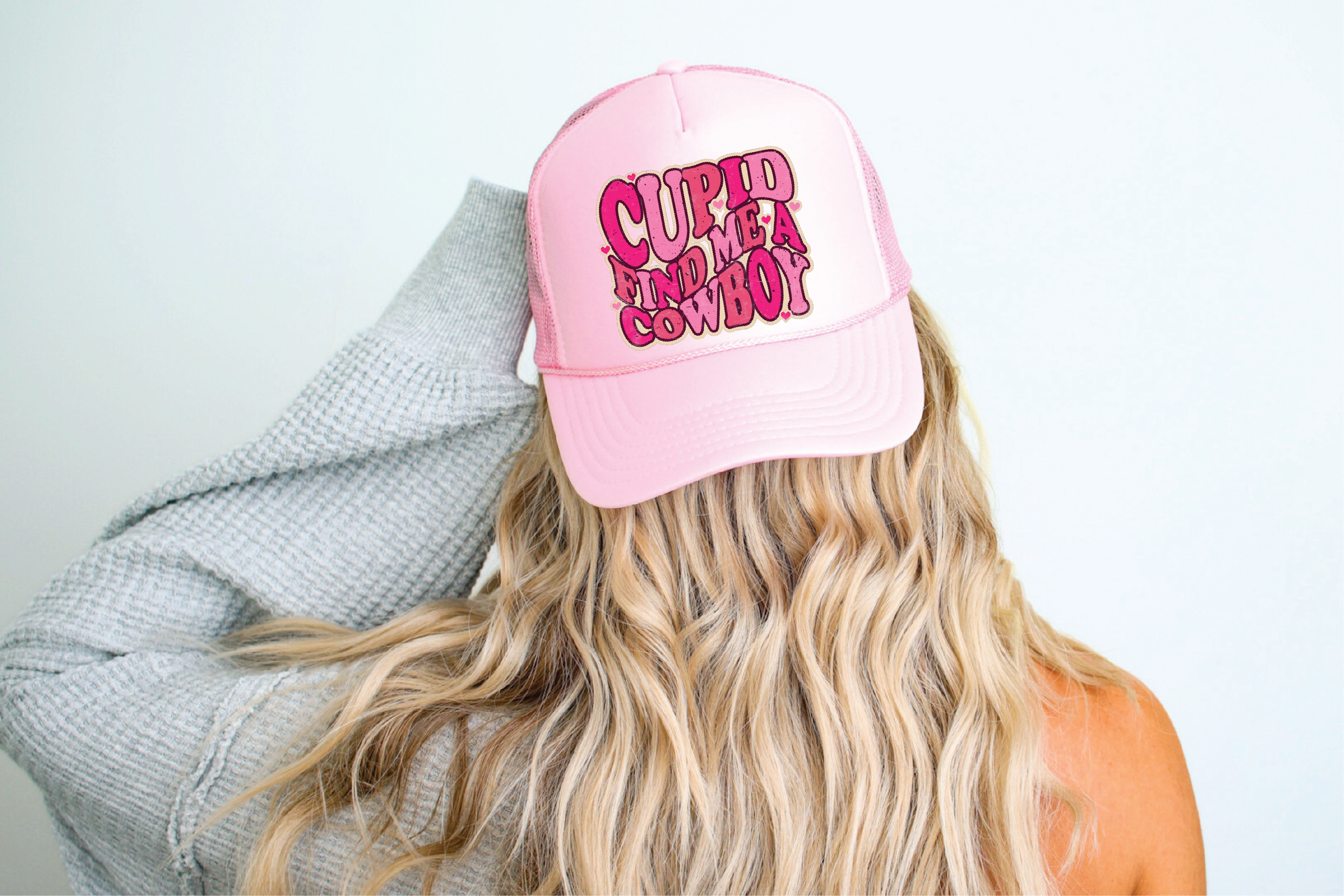 A person wearing a pink foam trucker hat with 'Cupid Find Me A Cowboy' printed in pink lettering on the front.