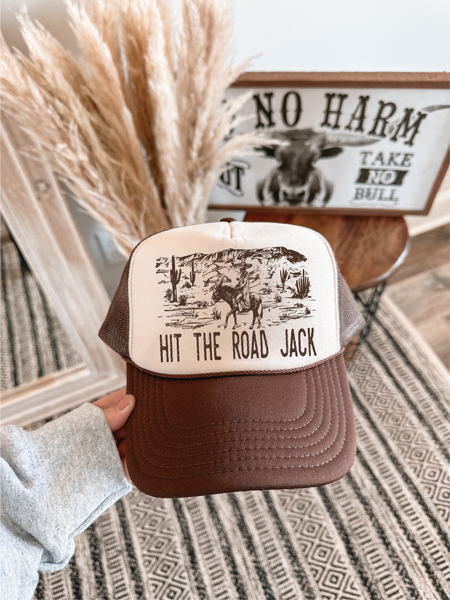A person holds a brown and cream trucker hat with a graphic of a cowboy riding a bucking horse and the text 'HIT THE ROAD JACK'. The background features a patterned rug, dried pampas grass, and a rustic wooden sign.