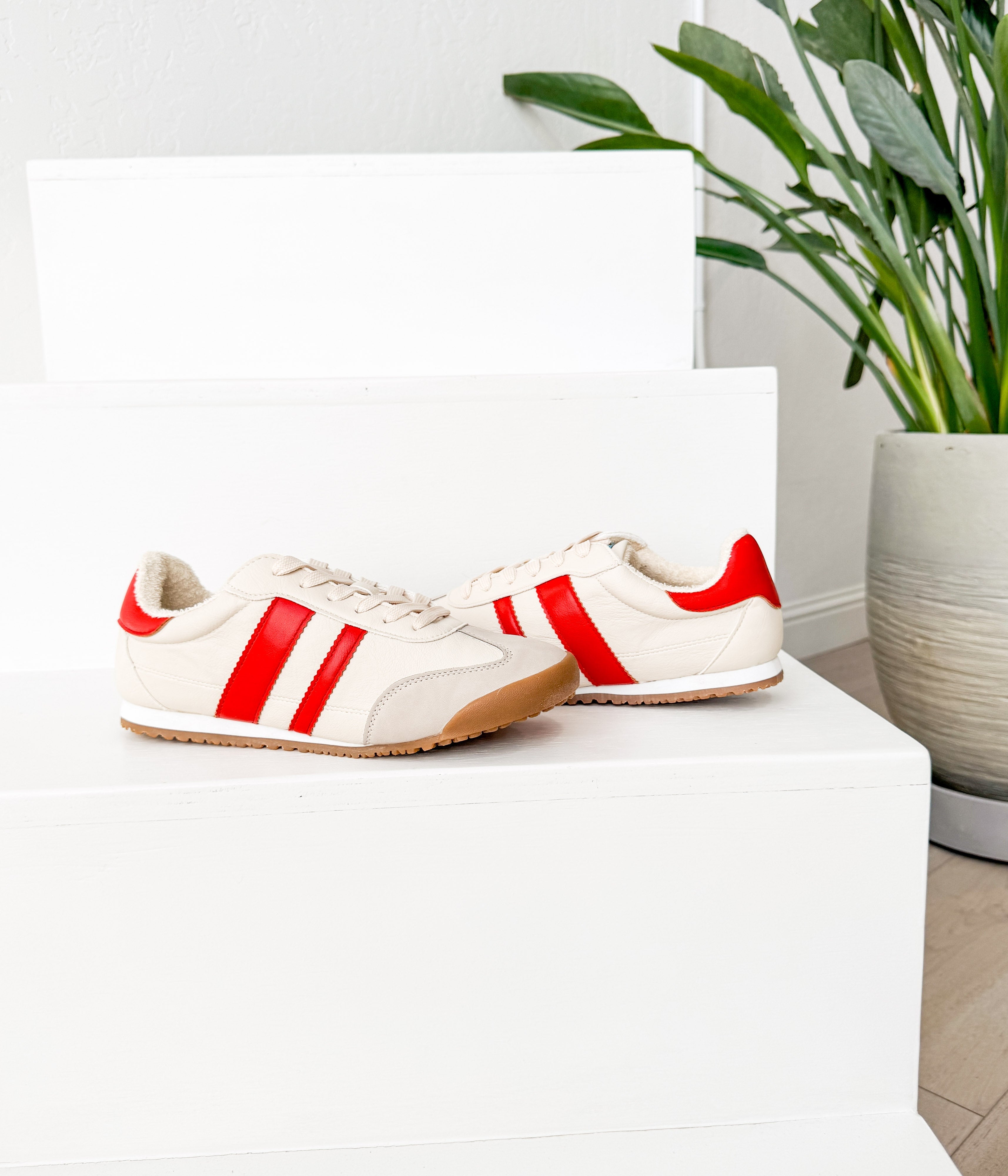 White sneakers with red stripes on a white surface next to a potted plant.