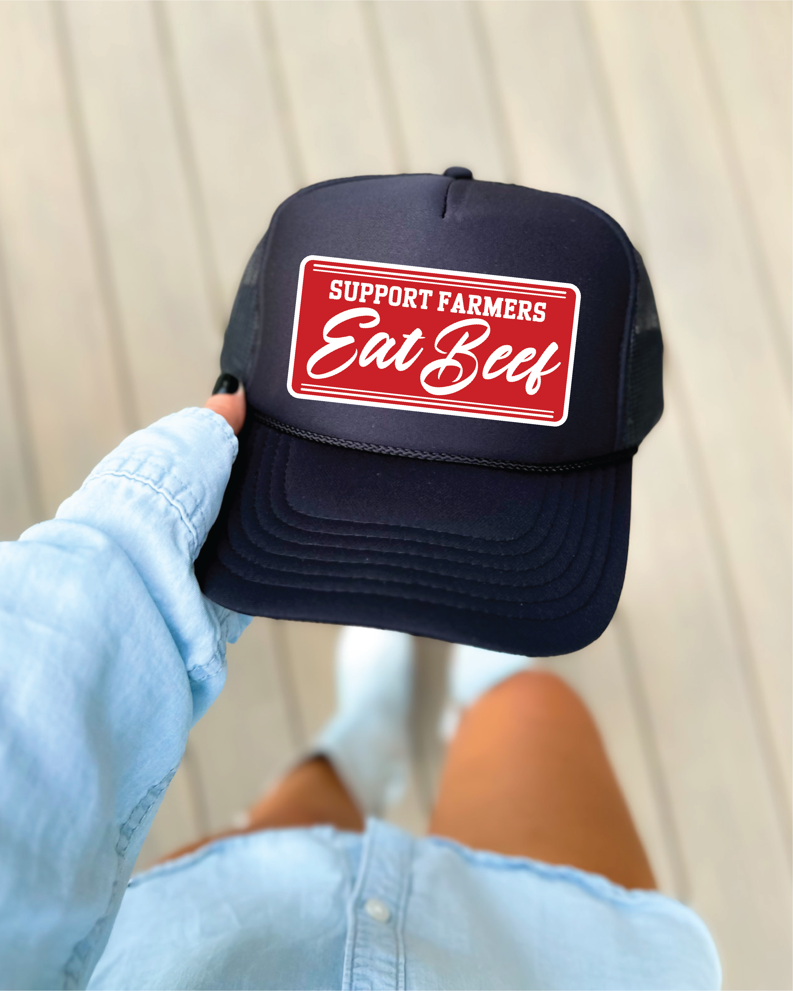 Navy blue cap with red and white 'Support Farmers Eat Beef' logo held by a person wearing a light blue jacket.